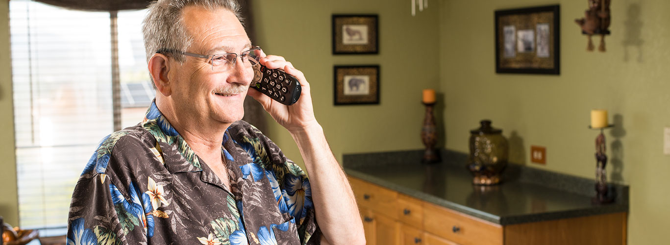 A middle-aged man, wearing glasses and a Hawaiian shirt, stands in front of a window talking on the phone in his home. There is a table behind him, which holds a vase and a candle. There is artwork on the wall behind him.
