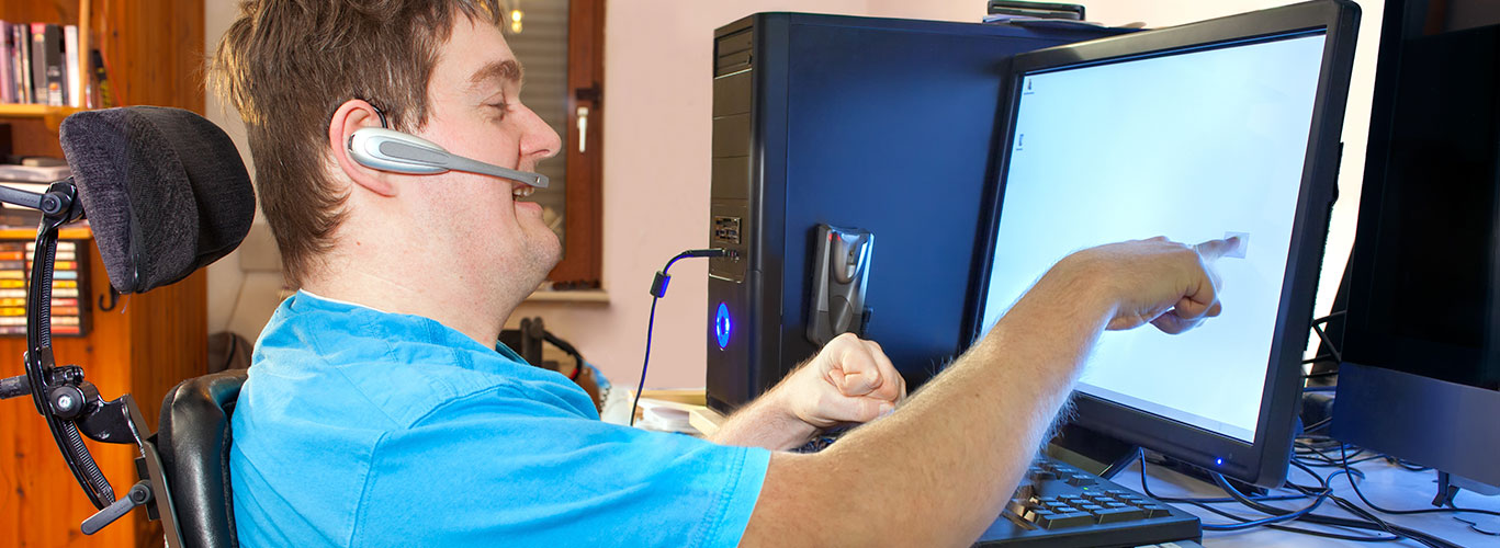 A middle-aged brunette man with a developmental disability sits in front of his computer at home, pointing at the screen. He is wearing an ear-piece to talk on the phone. There is a bookshelf behind him.