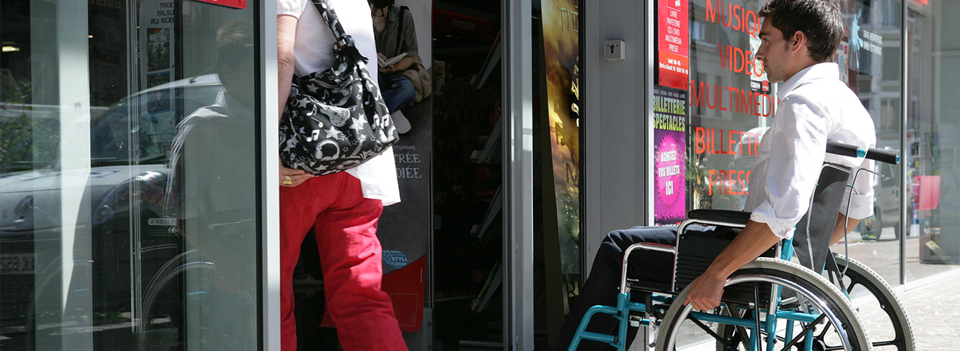 A young man in a wheelchair enters a store behind a woman walking through the glass door in front of him.