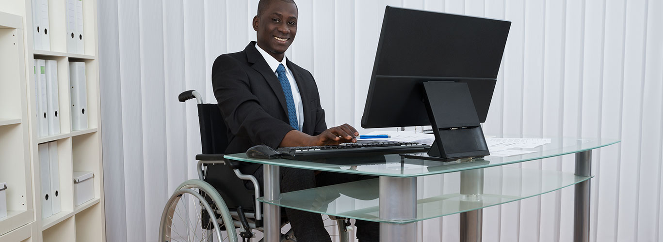 A young black man sitting in a wheelchair smiles while sitting at his glass desk holding a computer monitor at work. There is a white wall and white shelves filled with white binders surrounding him.
