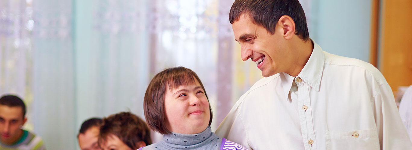 Close-up of young woman with a developmental disability, short hair wearing a purple and gray sweater smiling up at a taller man wearing a white shirt, blue jeans and a black belt.