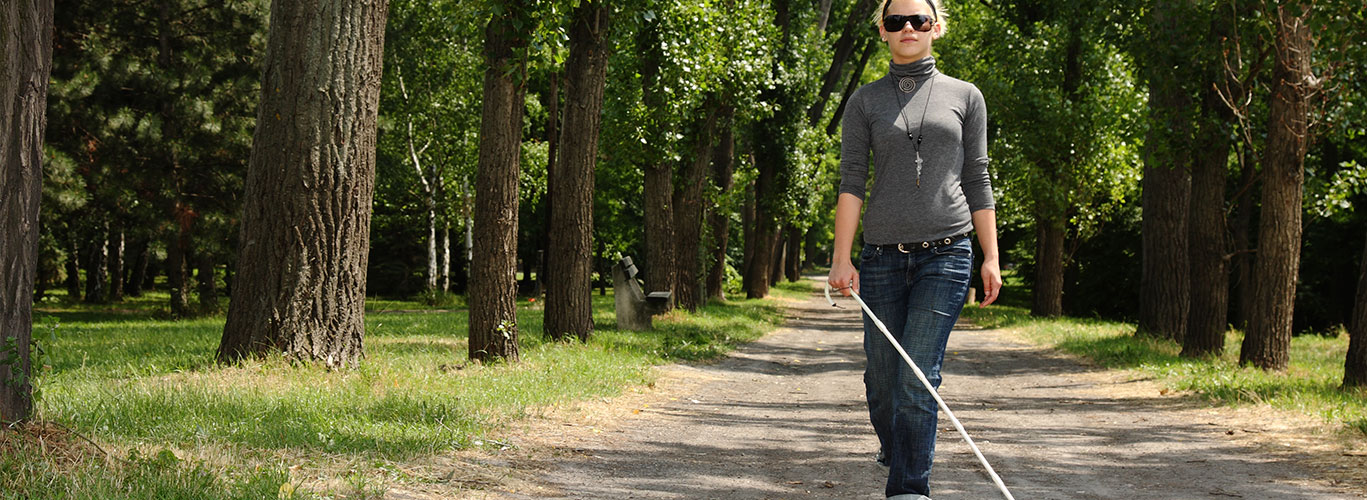 A young blonde woman wearing a turtleneck shirt, jeans, cross necklace and sunglasses walks alone on a path in a park with her white cane and dark glasses. She is flanked by trees and green grass.