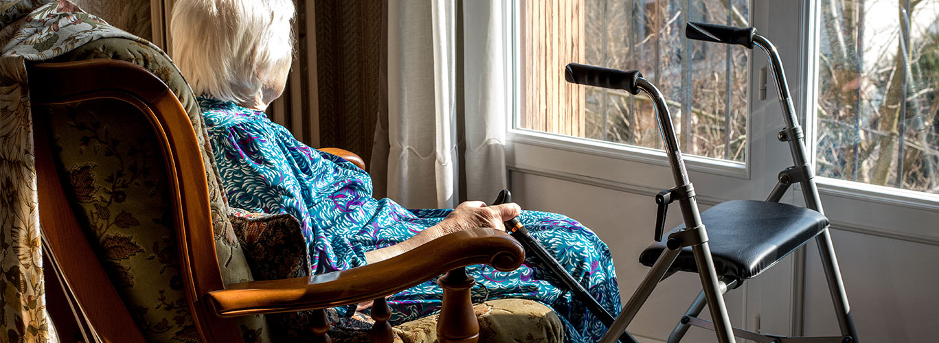 A senior woman sits in a rocking chair in her home looking out the window with her cane in one hand and her walker within reach. She is wearing a blue and purple abstract-patterned dress.