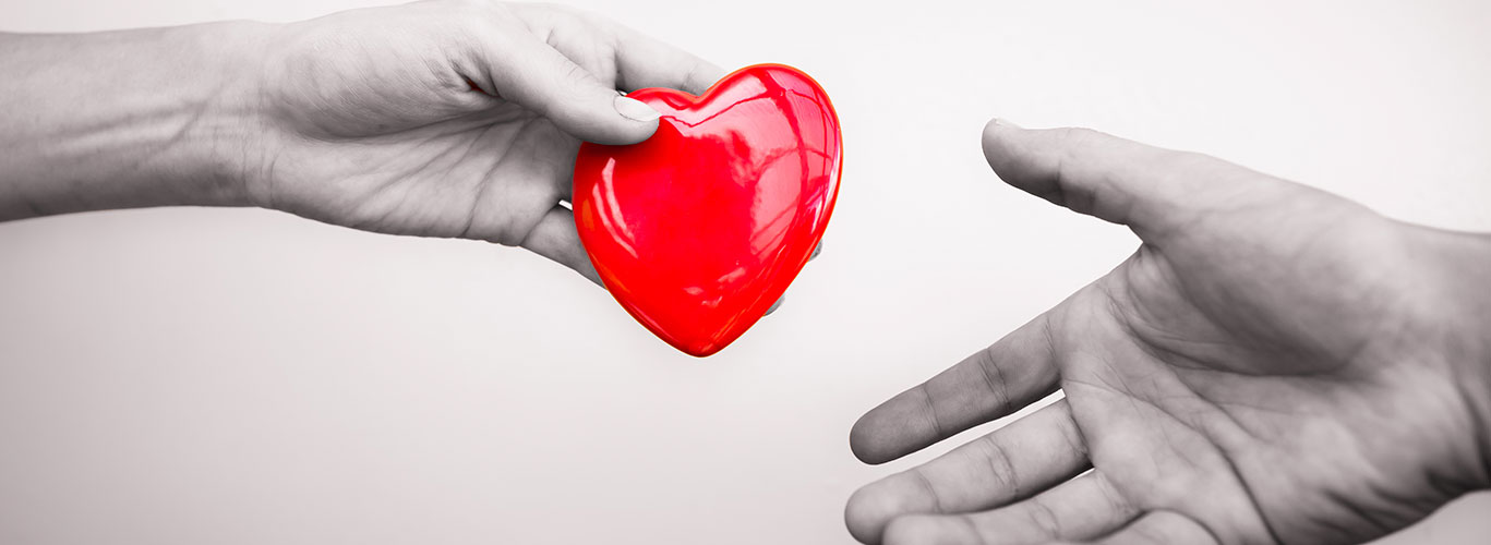 A black and white close-up photo of two hands stretching out toward each other. One is giving the other a plastic heart which is shown in color as red.