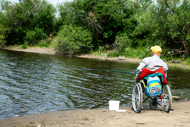 Man in a wheelchair fishing by the side of a lake.