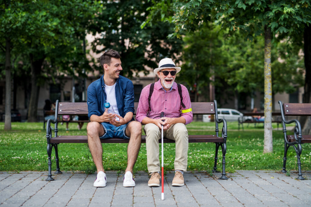 A young man and blind senior with white cane sitting on bench in park in city.