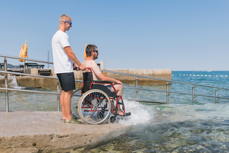 Two men at the beach, one in a wheelchair and the other standing behind him.